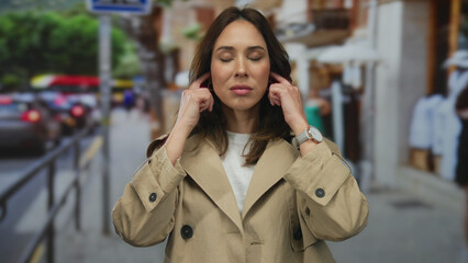 Woman in beige coat covers ears surrounded by busy city street to block out noise, demonstrating...