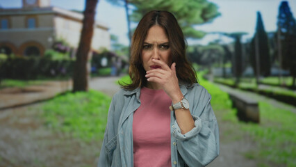 Fototapeta premium Young hispanic woman in a park expresses surprise and curiosity while enjoying a sunny outdoor setting, wearing casual attire and standing against a blurred green background.