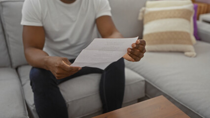 Obraz premium Young man reading a document in a cozy living room with soft lighting, emphasizing a relaxed home environment.