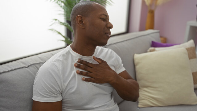 Young man experiencing discomfort sitting on sofa in living room, showing expression of pain, hand on chest, inside modern apartment with cushions and plants.