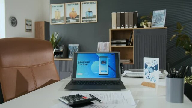 No people shot of laptop with app advertisement amidst paperwork, calculator, pens, and greeting card on desk in bank office