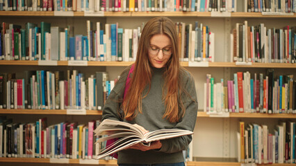 A student studies a book in the university library, standing between bookshelves with a backpack. A woman reading a book. 