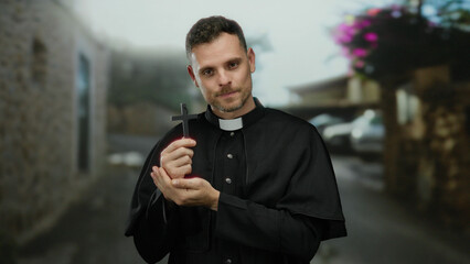 Priest holding cross outdoors wearing clerical clothing looking contemplative while standing on...