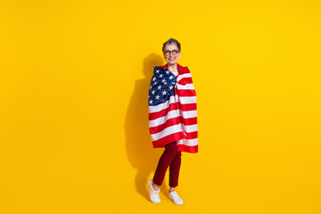 Cheerful senior woman wrapped in American flag stands against vibrant yellow background symbolizing...