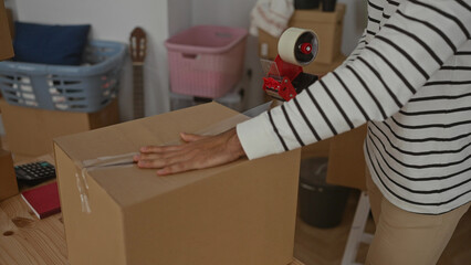 Fototapeta premium Man packing cardboard box in living room of new home with tape dispenser amid unpacked household items.
