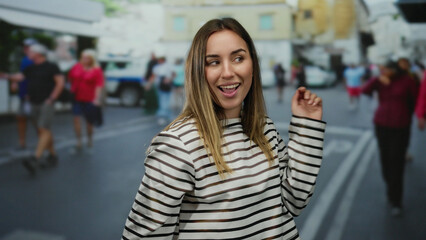Fototapeta premium Smiling young blonde woman stands on a lively city street wearing a striped shirt, capturing the essence of a cheerful outdoor urban environment.
