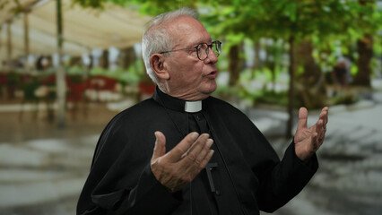 Senior priest man wearing glasses gestures outdoors on a street, with blurred green trees and...