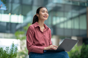 Fototapeta premium Business people thinking with laptop computer.Businesswomen outdoor working at green city background.Asia Business female sitting on big city urban street outside.City Streets With People concept 