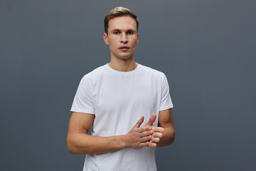 Handsome young man in plain white t-shirt stands against gray background looking serious and thoughtful, holding fingers together, close-up portrait, natural lighting, casual style, emotion concept