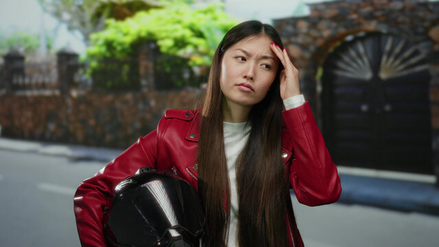 Woman in red jacket holding helmet on urban street appears thoughtful with serious expression against blurred city backdrop with stone wall and gate.