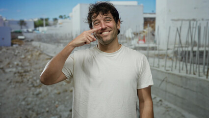 Fototapeta premium Hispanic man smiling in casual white shirt touches nose at construction site, showcasing a cheerful scene with urban and industrial background elements.