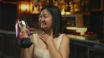 Chinese woman holding trophy smiling inside a warmly lit church with candles and religious decor highlighting her joyful expression and achievement.