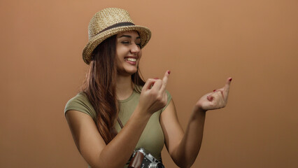 Hispanic woman smiling in a straw hat making a playful gesture against an isolated brown wall background, creating a vibrant and expressive mood.