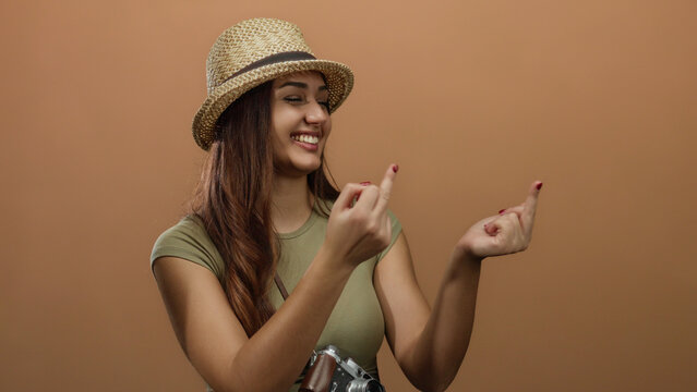 Hispanic woman smiling in a straw hat making a playful gesture against an isolated brown wall background, creating a vibrant and expressive mood.