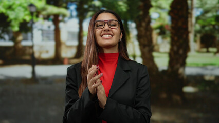 Woman clapping happily in a sunny park setting, wearing glasses and a vibrant red and black outfit,...