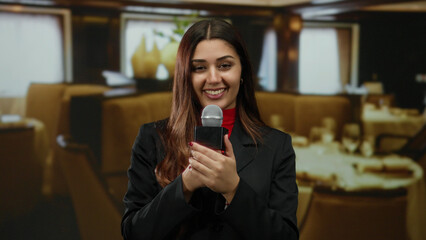 Young hispanic female reporter with microphone in elegant restaurant setting delivering news inside...