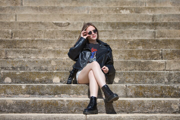pretty young blonde woman wearing sunglasses and dressed like a rocker is sitting cross-legged on...