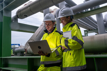 Two construction workers review plans on a laptop at an industrial site during the day near large...
