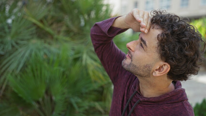 Young man looking up in urban park with greenery around, showcasing a handsome, hispanic adult amidst lush vegetation in city outdoors.