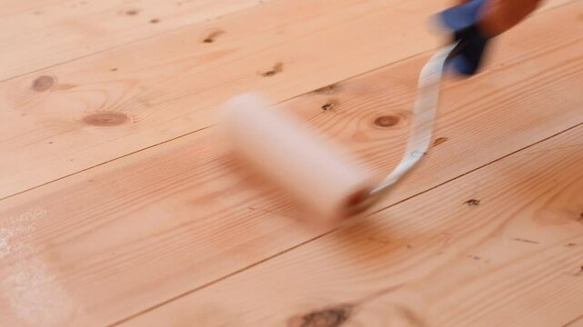 Applying transparent varnish onto wooden floorboards using a roller, creating a smooth finish and protecting natural wood texture.