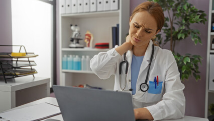 Woman doctor with stethoscope using laptop in clinic room while experiencing neck pain wearing white coat and thoughtful expression suggesting workplace stress.