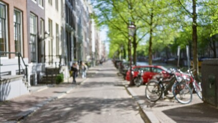 Blurred urban street scene in amsterdam featuring defocused canal, trees, and row of parked bicycles with people walking along the picturesque cobblestone pathway in daylight.