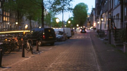 Blurred evening cityscape in a european town features bokeh streetlights, defocused trees, bicycles, cars, and cobblestone streets creating a serene urban ambiance.