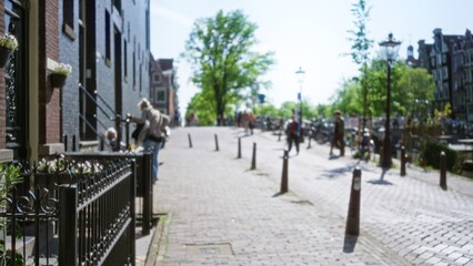 Blurred street scene capturing pedestrians and bicycles in a sunny european city setting with cobblestone road and traditional architecture amidst lush greenery.