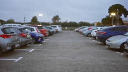 Defocused view of a parking lot filled with various colored cars under cloudy skies, showcasing a soft bokeh effect with blurred background and scattered lights.