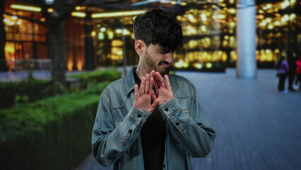 Young man with a disgusted expression standing outdoors on a city street in a denim jacket during...