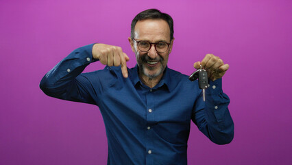Elderly man smiling and pointing at car keys against pink background wearing glasses and blue shirt