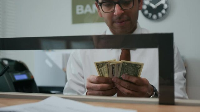 Close-up shot of man in glasses counting, holding stacks of American dollar bills in his hands behind teller window in bank office