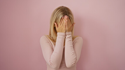 Woman covering face with hands while standing against a soft pink background, conveying the concept...