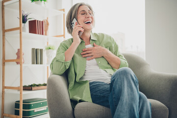 Mature woman laughing while talking on mobile phone, sitting comfortably in an armchair at home interior
