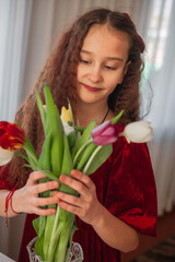 Naklejka premium Smiling girl in a red velvet dress arranging colorful tulips in a vase by the window, surrounded by soft daylight and spring atmosphere.