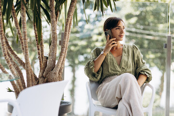 Fototapeta na wymiar Young woman sitting on a chair under palm trees and talking on the phone