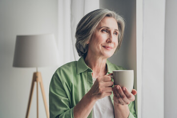 Mature woman enjoying leisure at home while standing by a sunny window and holding a warm cup in...