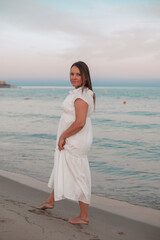 Smiling woman in a white summer dress walking barefoot along the seashore at sunset. Gentle waves and pastel sky create a warm, relaxing vacation mood by the sea.