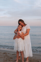 Mother and daughter in matching white dresses hugging on the beach at sunset. Peaceful sea and soft pastel sky in the background. Family love and tenderness.