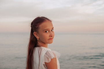 Back view of a young girl in a white dress turning her head toward the camera on a beach at sunset....
