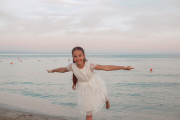 Joyful girl in a white dress playing on the beach, balancing on one leg with arms outstretched like...