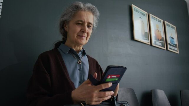 Low angle shot of elderly woman with gray hair smiling while using her smartphone in waiting room of modern bank office