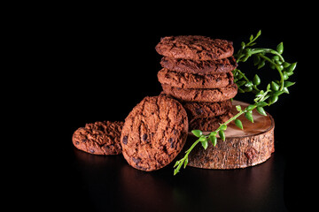 Chocolate chip cookies stacked on wooden base with decorative green leaves and dark background, suitable for advertising and promotion.