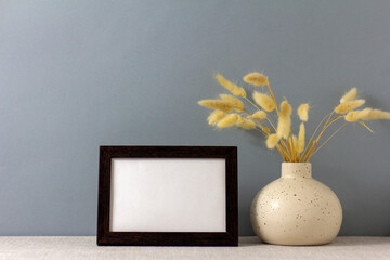 The layout of an empty photo frame on the table. A vase with dry grass. A minimalistic workspace. A place to copy.