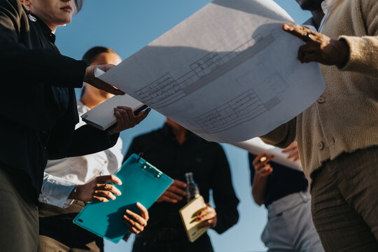 Business people collaborate under clear skies, analyzing architectural blueprints and documents. The image captures teamwork, planning, and outdoor discussions in a corporate setting.