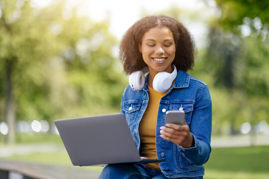 Happy Black student woman using smartphone and laptop outdoors, smiling african american female multitasking outside in green park, enjoying modern education, copy space - Powered by Adobe
