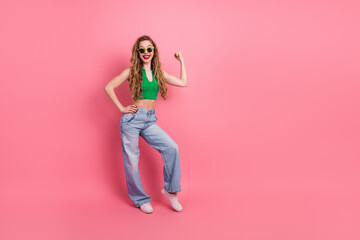 Fashionable young woman in a green top and jeans posing confidently against a vibrant pink studio...