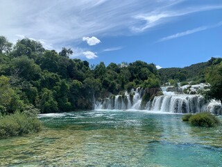 Fototapeta premium Stunning turquoise river with cascading waterfalls surrounded by lush green forest under a bright blue sky in Plitvice National Park