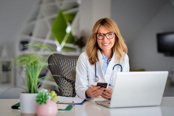 Mature healthcare worker sitting at doctor's office and using phone