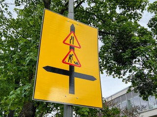 Bright yellow temporary road sign displays merging lanes, road work symbols, and directional arrows...
